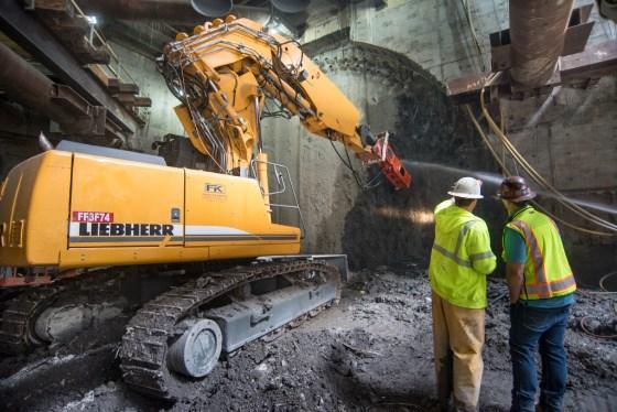Liebherr excavator demolishes concrete wall inside underground station cavern while two workers in safety vests observe.
