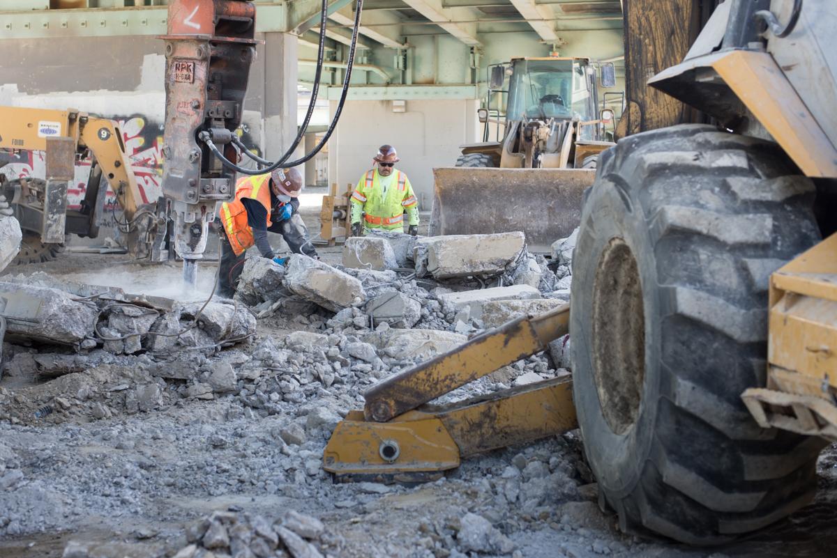 Workers in safety vests operate heavy machinery to break up concrete and rebar in a construction work yard.
