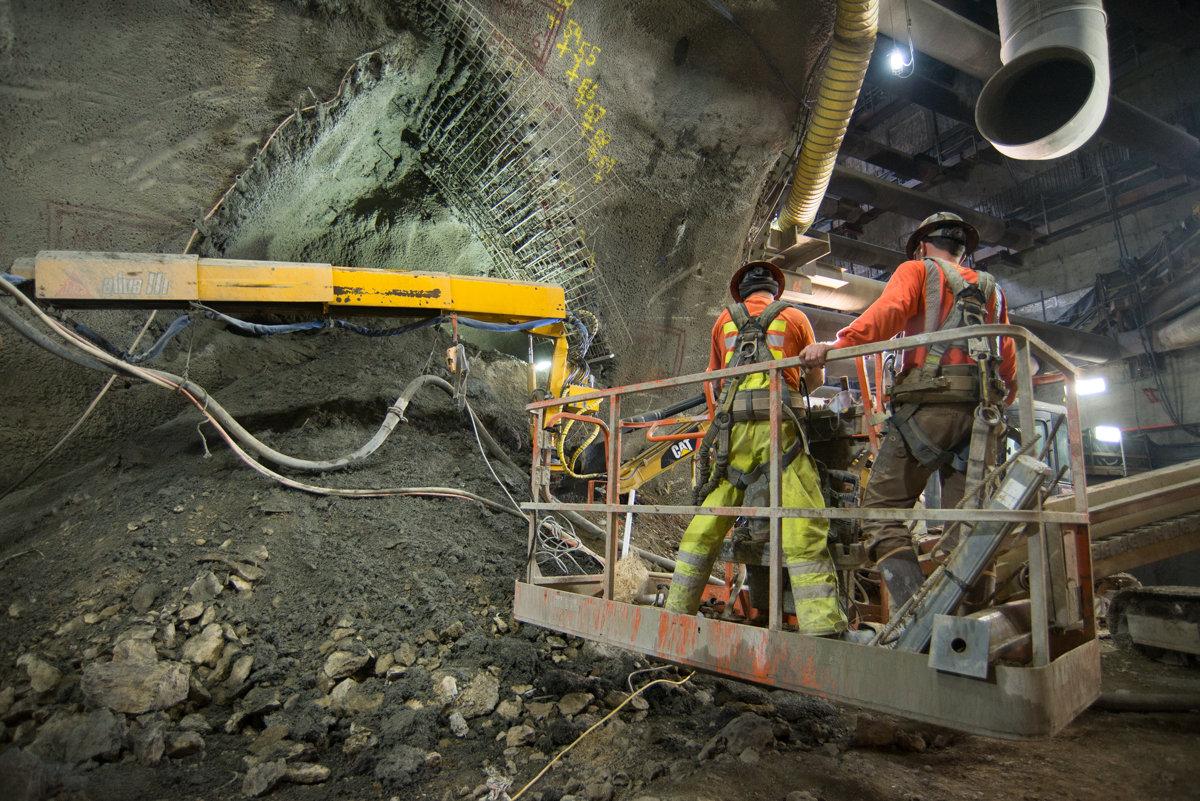 Workers in safety gear on elevated platform near underground excavation site with drilling equipment.