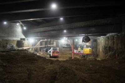 Underground construction site with heavy equipment and lighting beneath concrete structure.