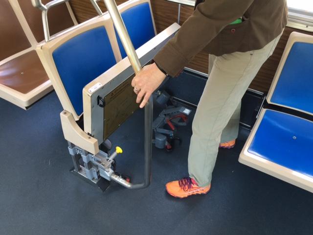 Person demonstrating how to unlock and fold up a transit bus seat by pulling the yellow knob on the stanchion.
