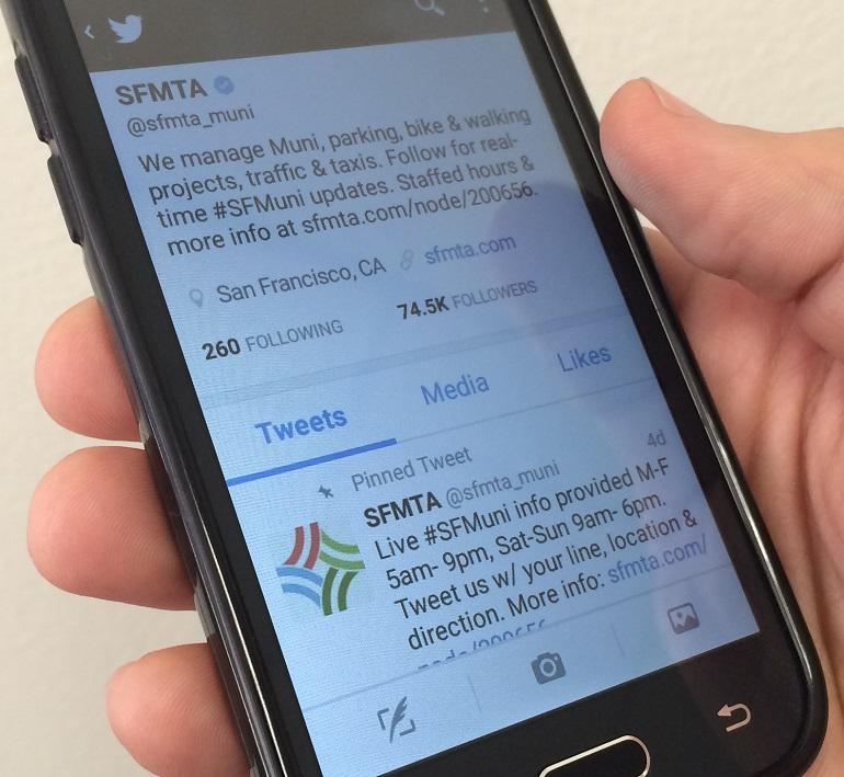 Hand holding smartphone displaying SFMTA's Twitter profile page with 74.5K followers and pinned tweet about Muni service hours.
