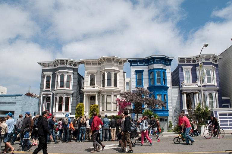 Pedestrians and cyclists on Valencia Street in front of colorful Edwardian Victorian homes during Sunday Streets event.