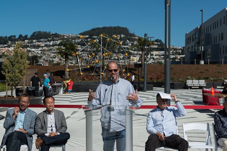 Man speaking at podium during Unity Plaza ribbon cutting ceremony with seated attendees and yellow playground structure visible behind.