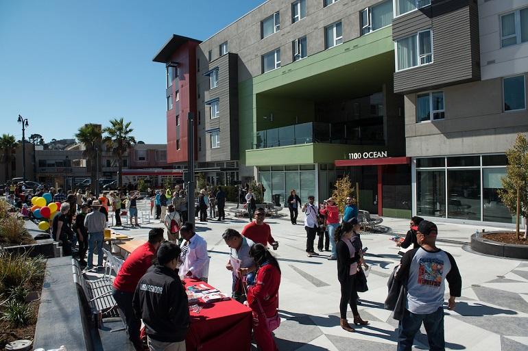 Crowd of people gathered at Unity Plaza during a community celebration, with modern mixed-use buildings featuring red and green architectural elements in the background.