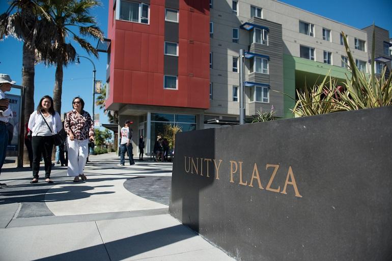 Unity Plaza sign on a low wall with pedestrians walking past colorful residential buildings on a sunny day.