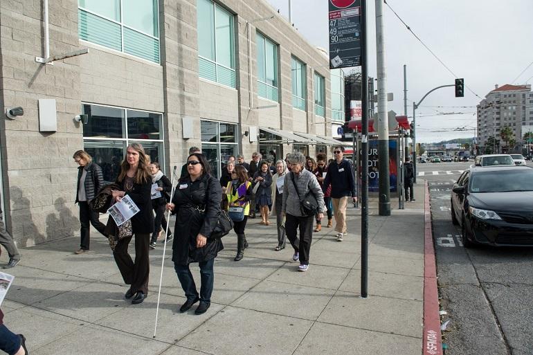 Group of people walking on Van Ness Avenue sidewalk during a tour, with buildings and street infrastructure visible.