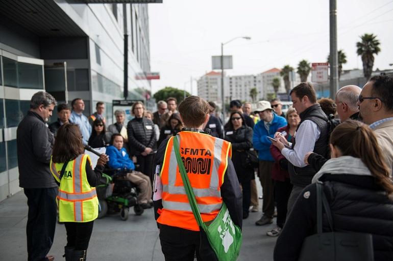 SFMTA staff member in orange safety vest addresses crowd of approximately 40 people outside SFMTA headquarters on Van Ness Avenue.