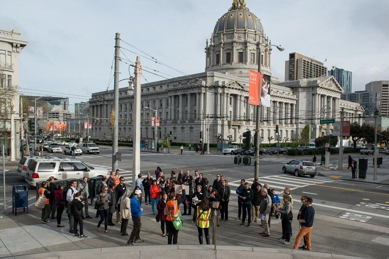 Tour group gathered at Van Ness Avenue and McAllister Street with San Francisco City Hall dome visible in background.