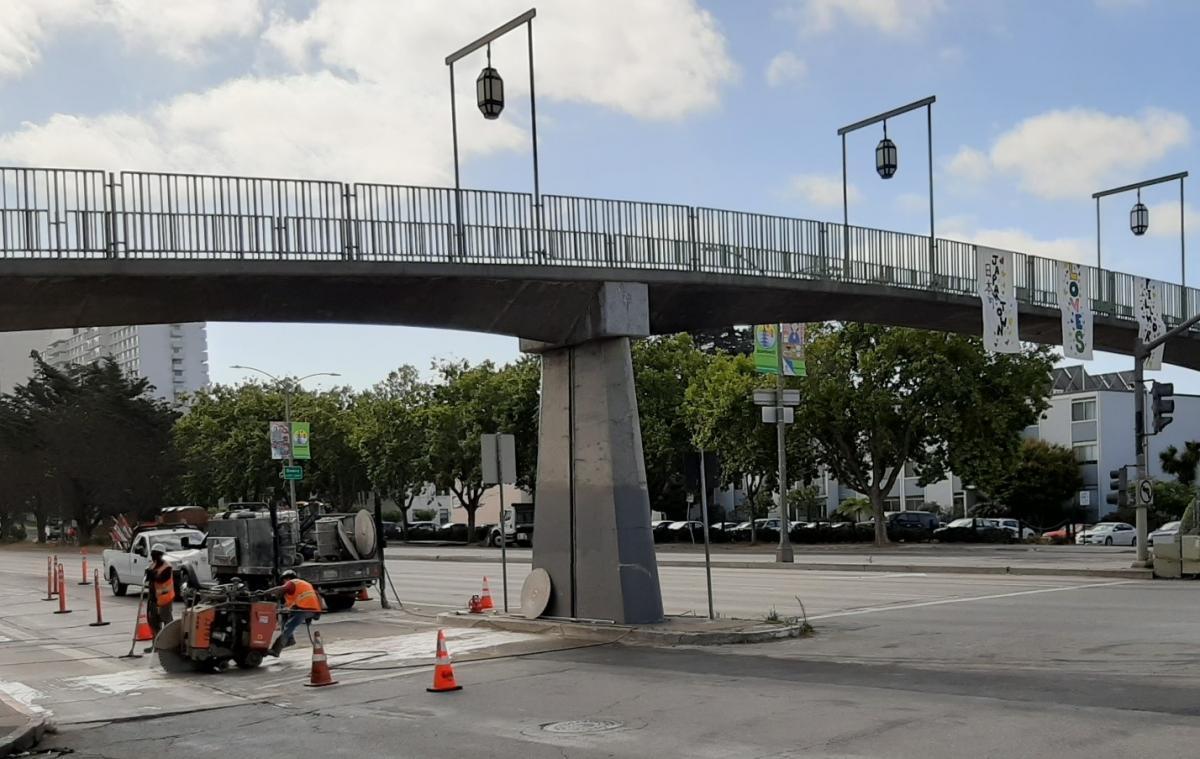 Photo: Crew saw-cutting for a new fire hydrant pipe at Webster and Geary, with banners on the pedestrian bridge in the background “Japantown Loves Fillmore”