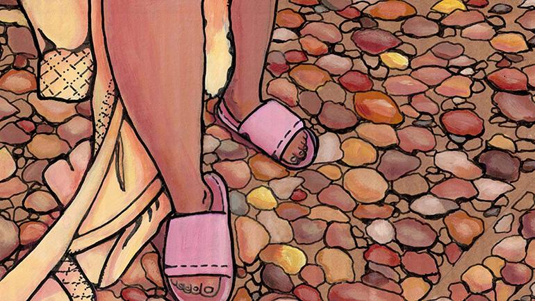 Legs draped in a robe and feet in open-toed slippers walk across a surface of small stones in varying sizes and brownish, yellowish and reddish colors.