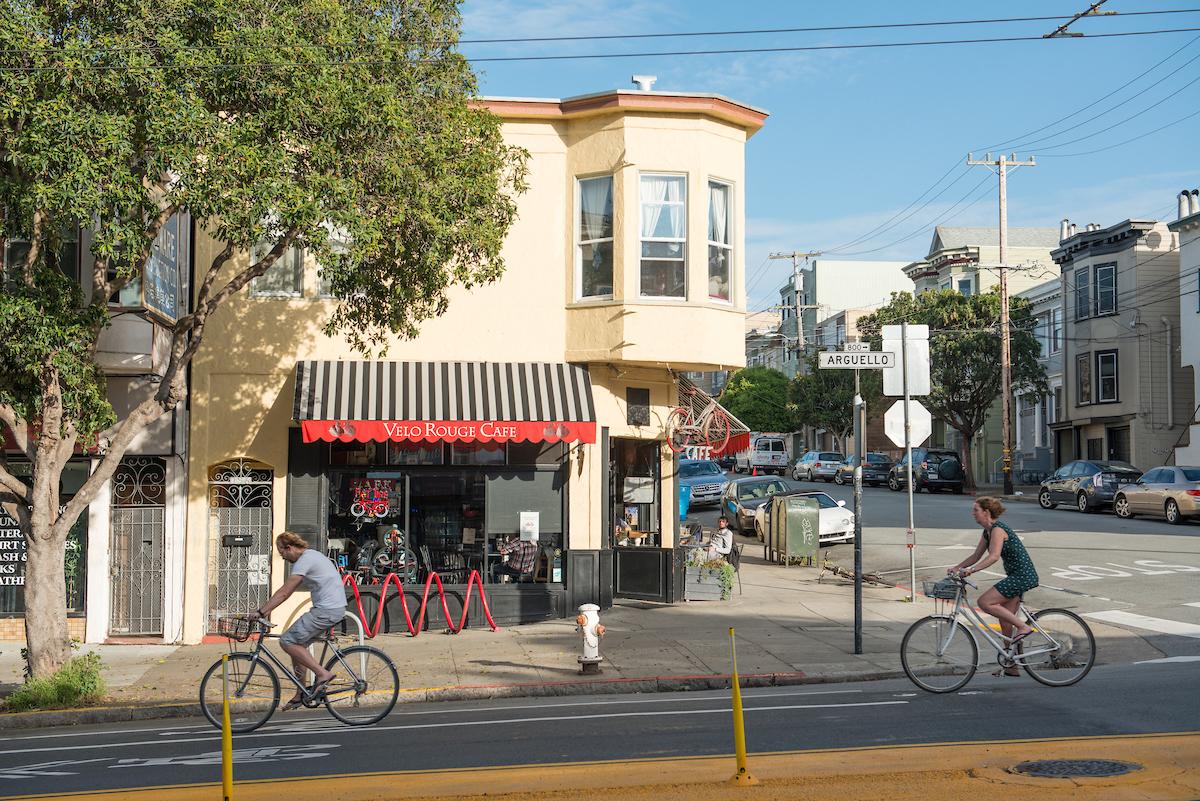 Two people ride bikes on the street in front of a restaurant with people sitting outside.