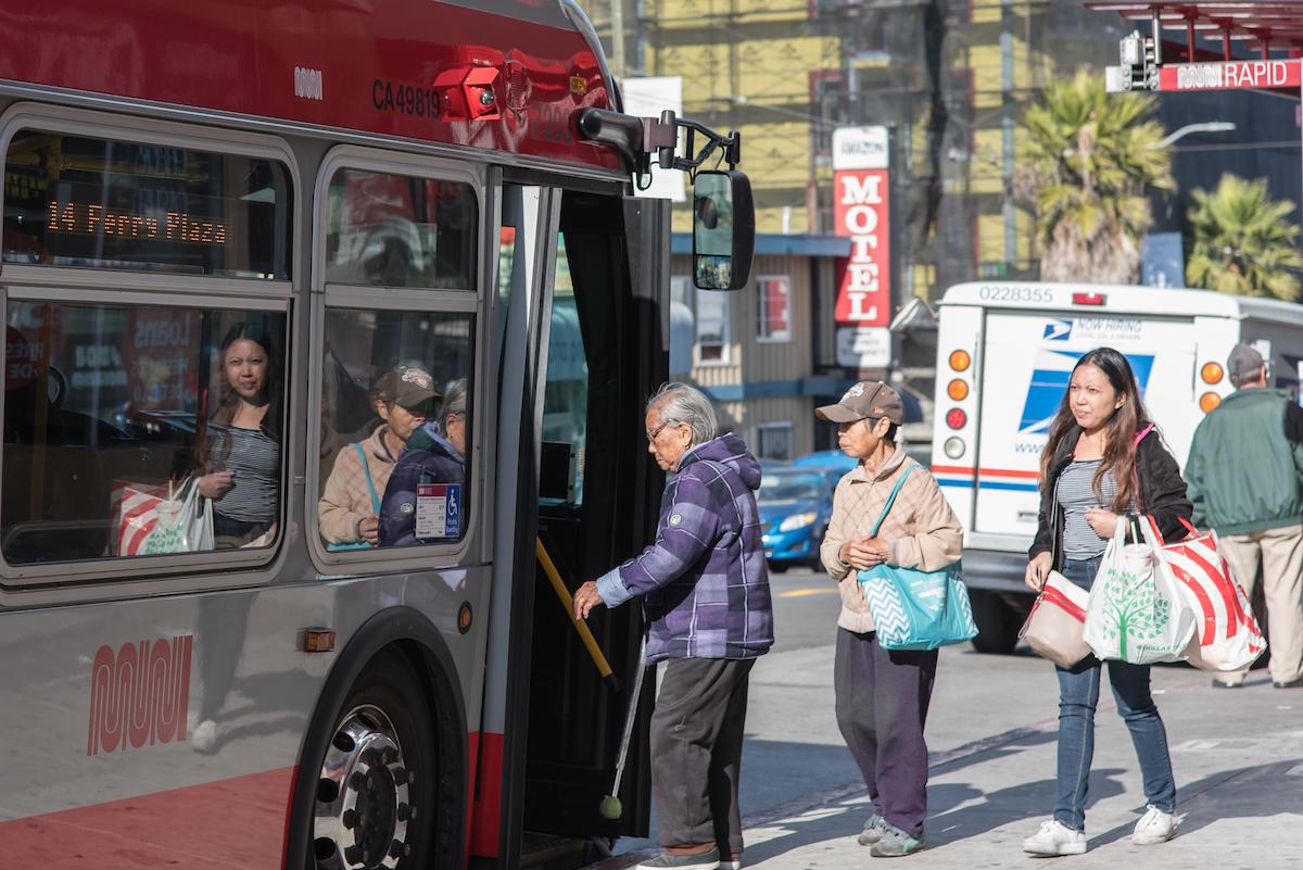 A senior and two women carrying shopping bags wait in line to board a 14. We see other vehicles moving in the background.