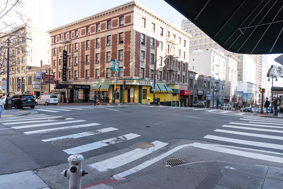 An intersection in San Francisco with crosswalks, surrounding tall buildings and trees as people walk towads the intersection.