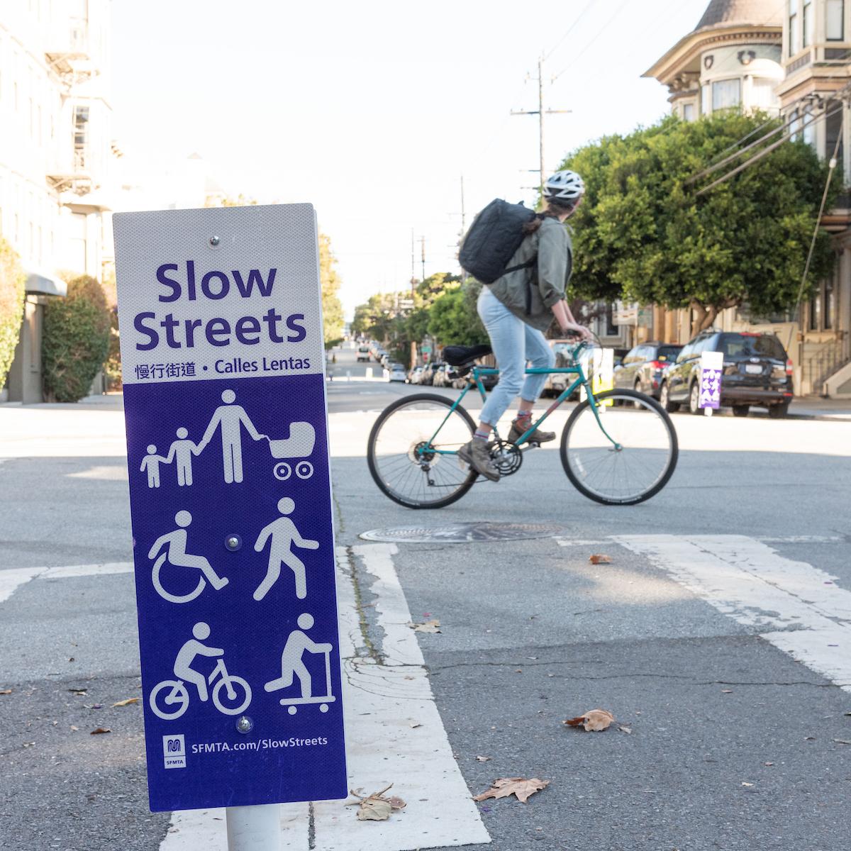 Close up image of a street sign with the words "Slow Streets." Under white figurine wheel users and pedestrians are seen. There is a bicyclist in the background