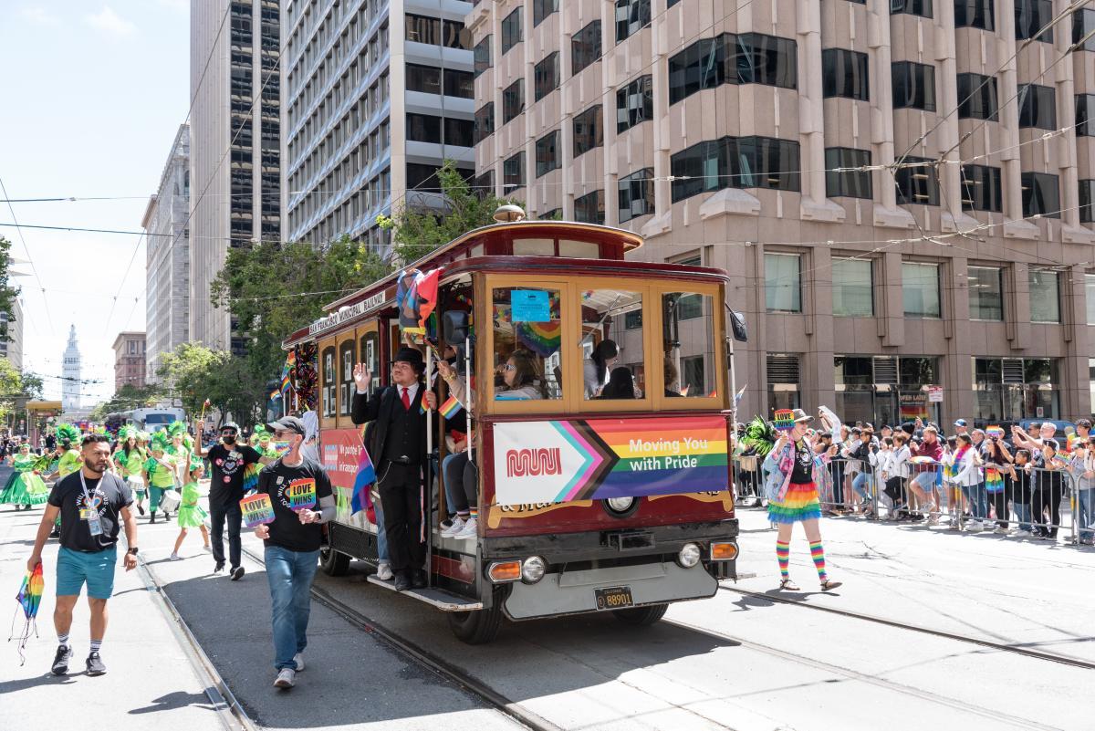 People march at a parade on the street with rainbow flags around a historic cable car.