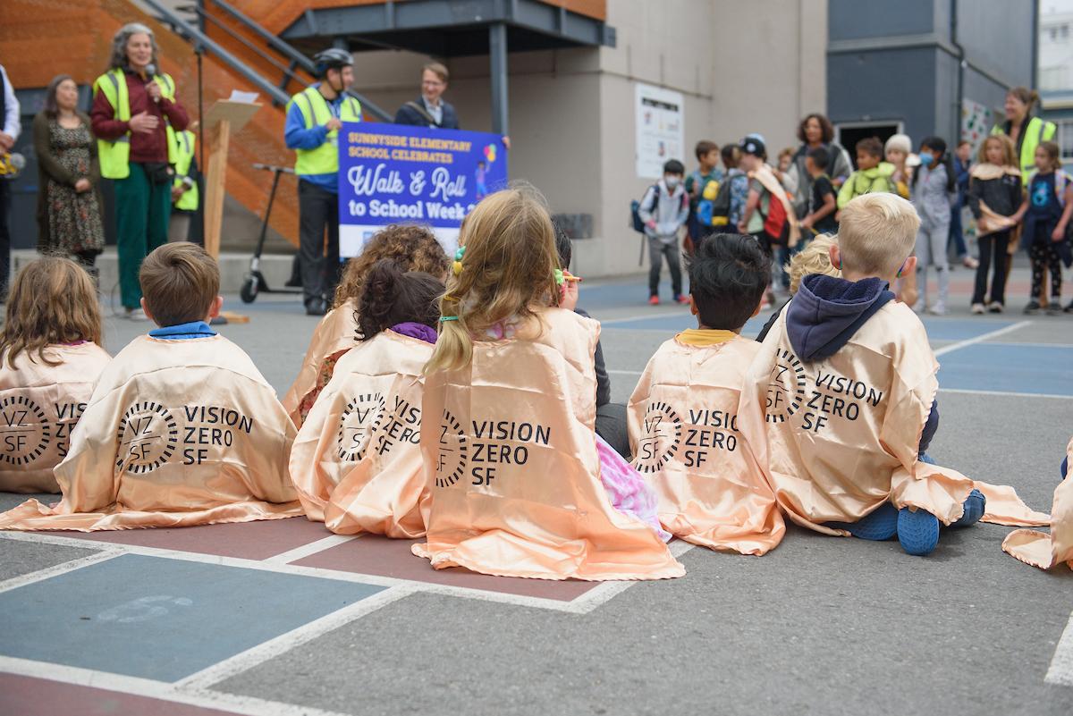 Children on a playground listening to adults, some holding a sign.