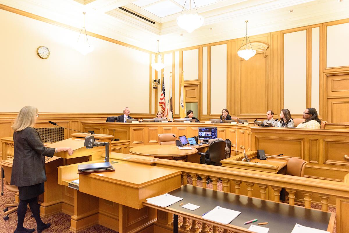 A meeting held in a building with brown furnishings with a person presenting to a group of people sitting at a semi-circular table.