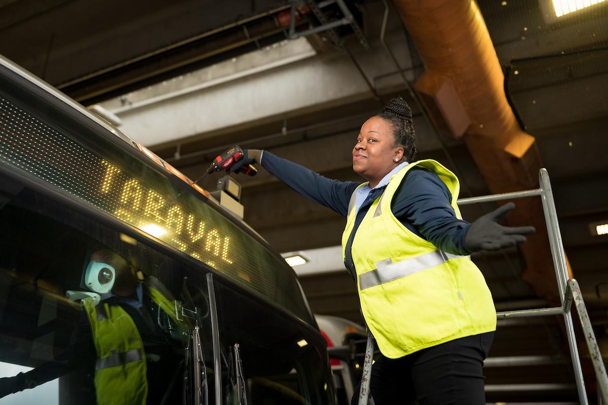 Nicole Humphrey changes a clearance light on a Muni bus. We see her with a drill, standing by the top of the bus.