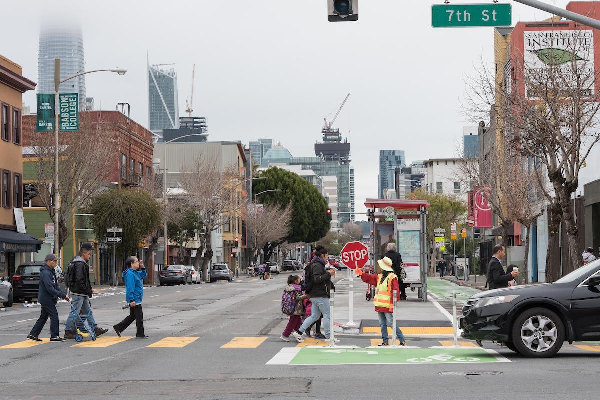 Seniors, children and others cross 7th Street at Folsom with help from a crossing guard who stands in the middle of the crosswalk as a car approaches.