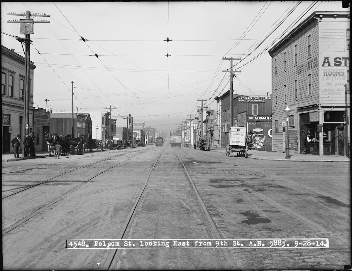 Black and white photo of Folsom Street in wide view, lined by commercial buildings with horses and carriages parked out front.