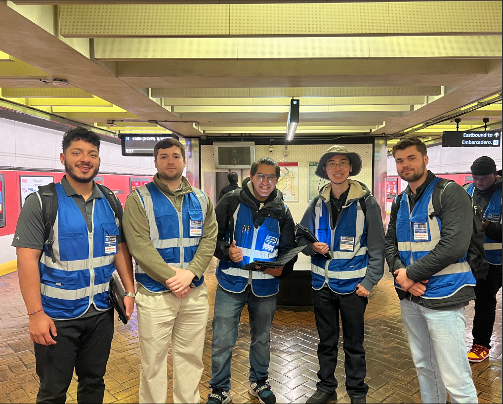 Survey takers wearing blue vests and badges smile as a group as they stand on a Muni Metro platform.