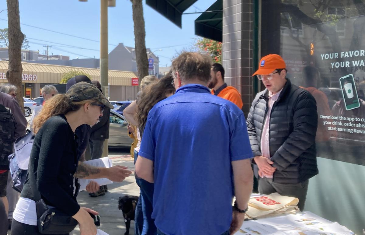Several people stand in front of a table where a man in an SFMTA hat is speaking as a street corner.