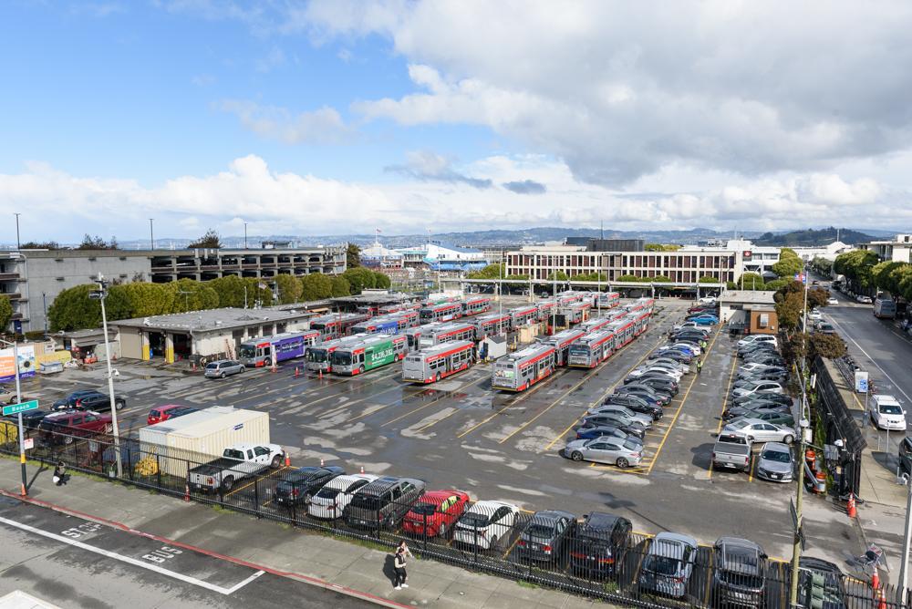 Kirkland Yard full of buses with parked cars lining the sides of the yard. People pass by on the adjacent sidewalk.
