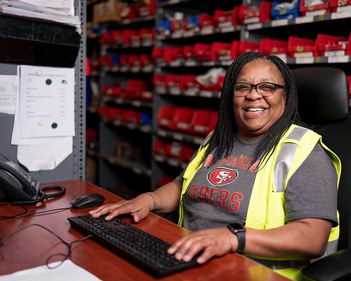 Cotton smiles from her desk chair with her hands on the keyboard. We see shelves full of parts behind her.
