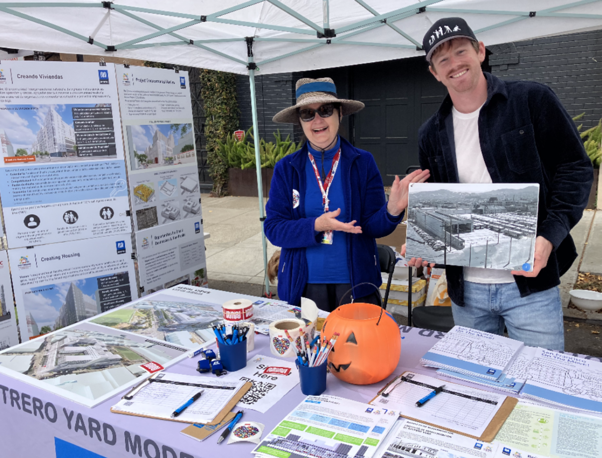 Two people looking at the camera while smiling and gesturing towards visual and printed information on a table