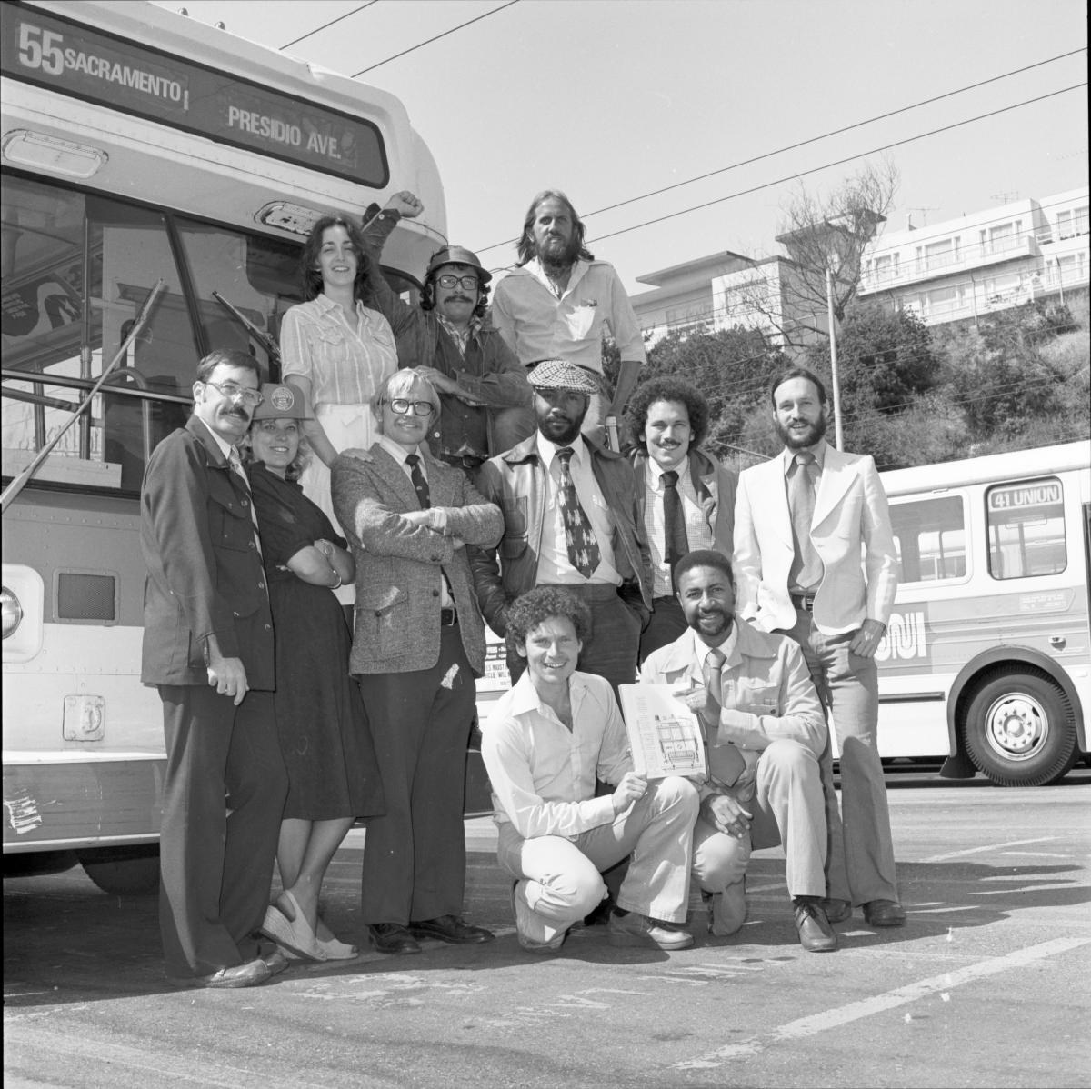 A dozen people dressed in business casual posting for a group photo in front of a bus in what seems to be a bus yard.  They are positioned in several different tiers. 