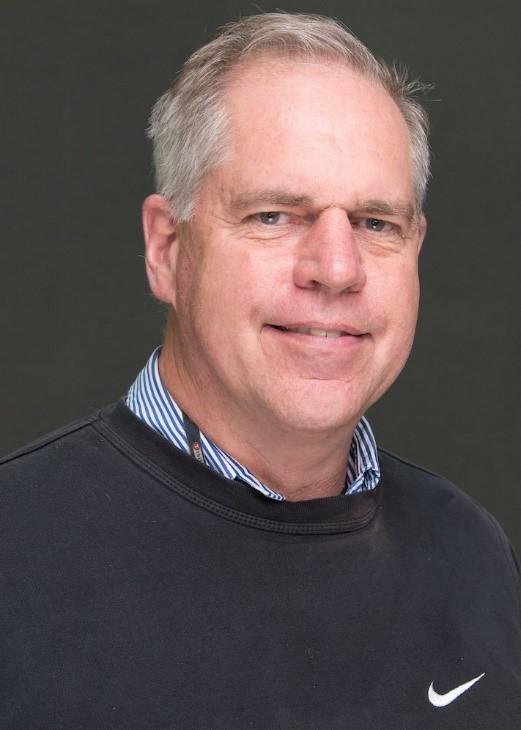 Man in front of grey background wearing blue sweatshirt and blue striped shirt.