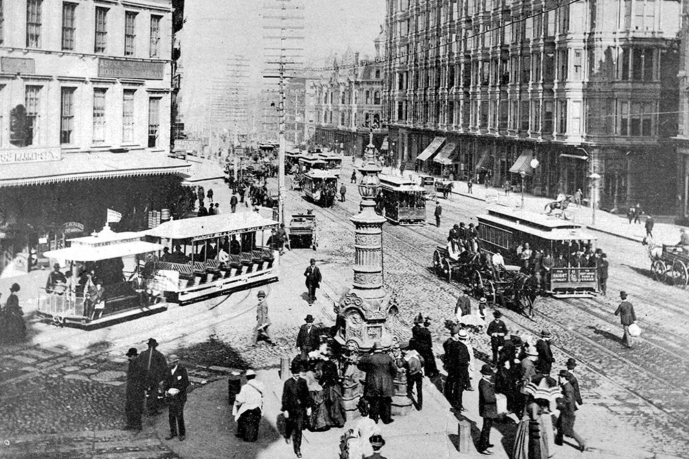 Black and white photo of intersection showing people and cable cars on streets. 