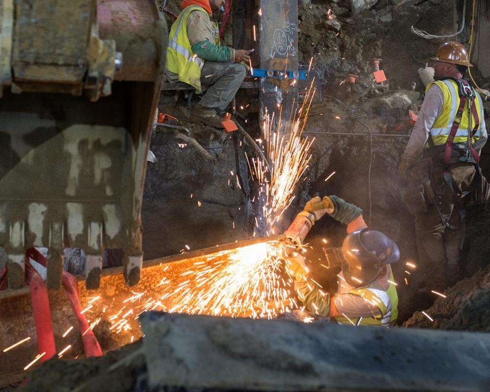 A metalworker cleans up the ends of a large steel I-beam for welding, which will be used as a bracket at the south headwall.