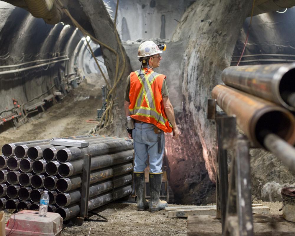 Drill casings surround a worker pausing for a bulldozer to pass during excavation of the Chinatown Station platform cavern.