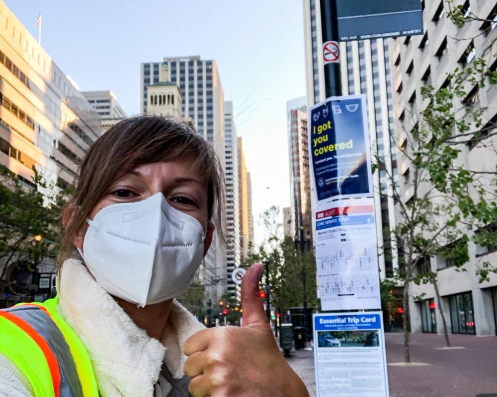 Photo of SFMTA staff giving a thumbs up after posting face mask, wayfinding, and ETC signs at a bus stop on Market Street.