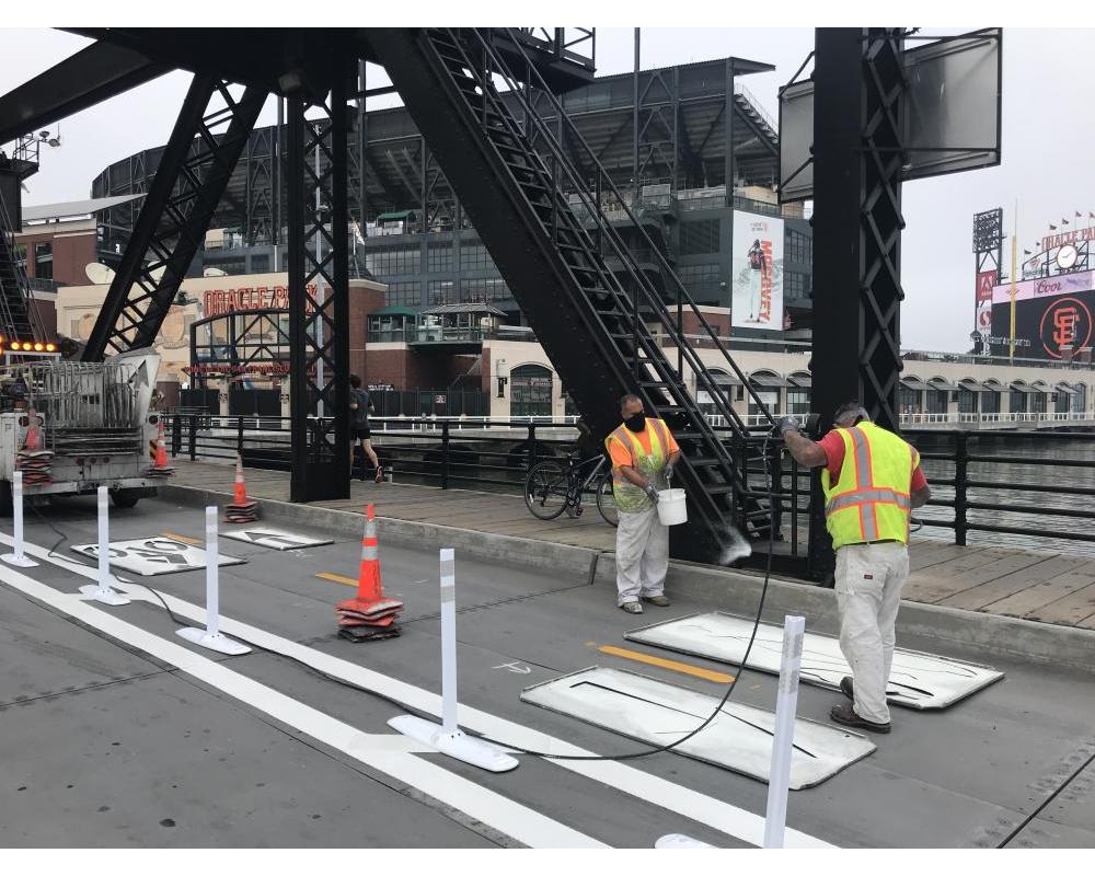 two people wearing yellow safety vests spray painting white paint using stencils in a two-way bikeway with plastic delineators