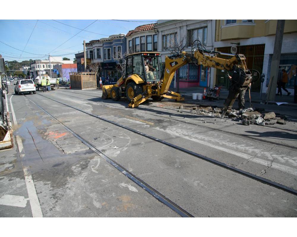Excavator with extended arm and alongside the rail tracks and residential homes.
