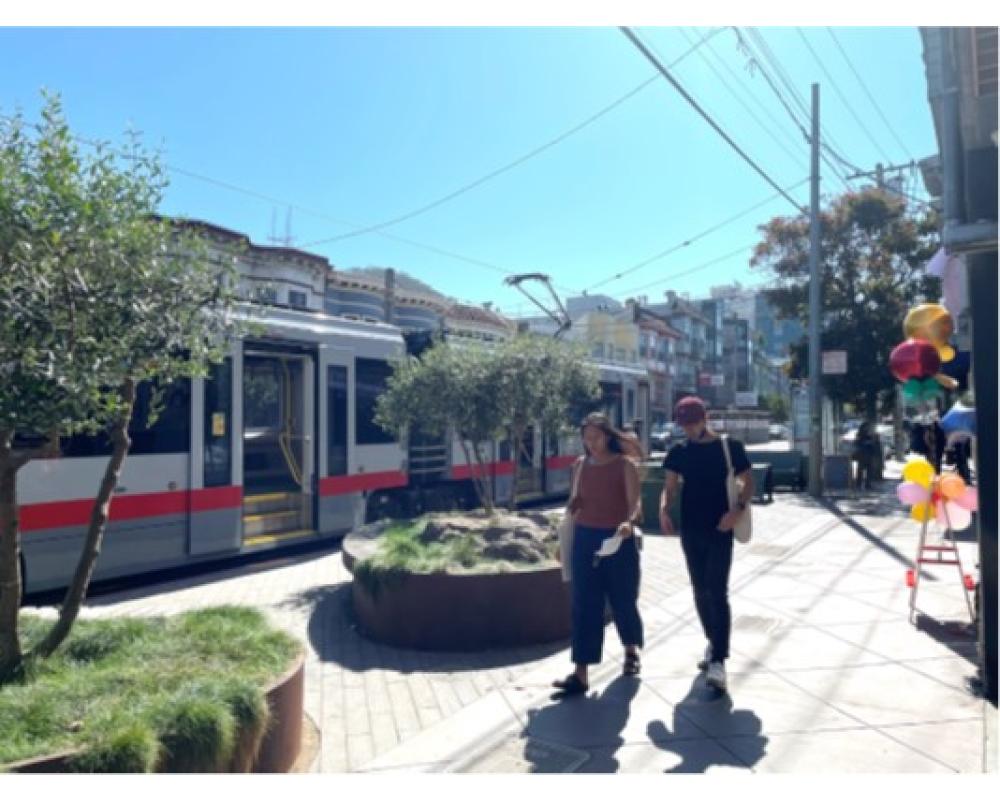People walking by sidewalk planters near train. 