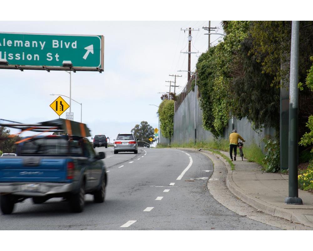 Bicyclist walking their bike on the sidewalk westbound on Alemany Blvd.