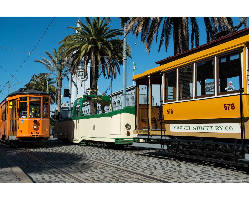 An orange historic streetcar passes a green and cream open-air Boat Tram and another orange historic streetcar. Palm trees and Ferry Building in the background.
