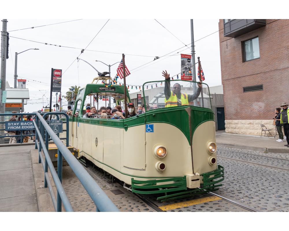 Cream and green open-air streetcar, a Boat Tram, drives near the Embarcadero. The operator waves.
