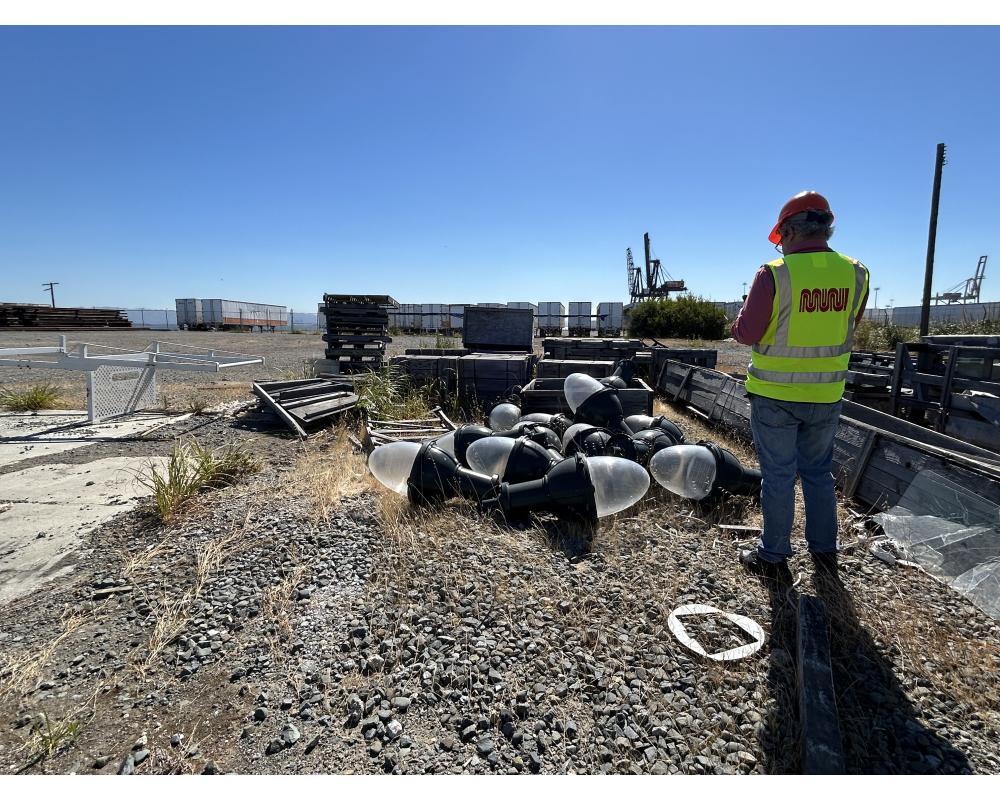 Gravel storage yard with shrubbery. At SFMTA employee in a yellow safety vest looks down at large lightbulbs and bulky material being stored
