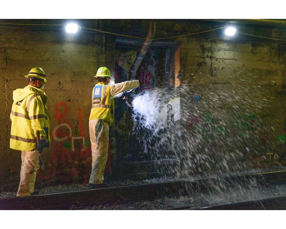Two crew members wear yellow safety gear inside a Muni Metro tunnel as they flush standpipes for Fix-It Week.