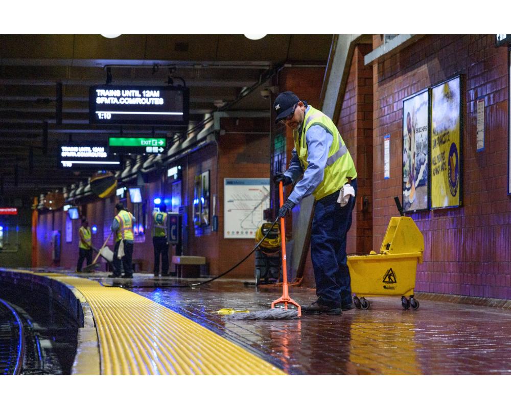 Custodian wearing a yellow vest and blue uniform mops the floor of a Muni Metro station as part of Fix-It Week.