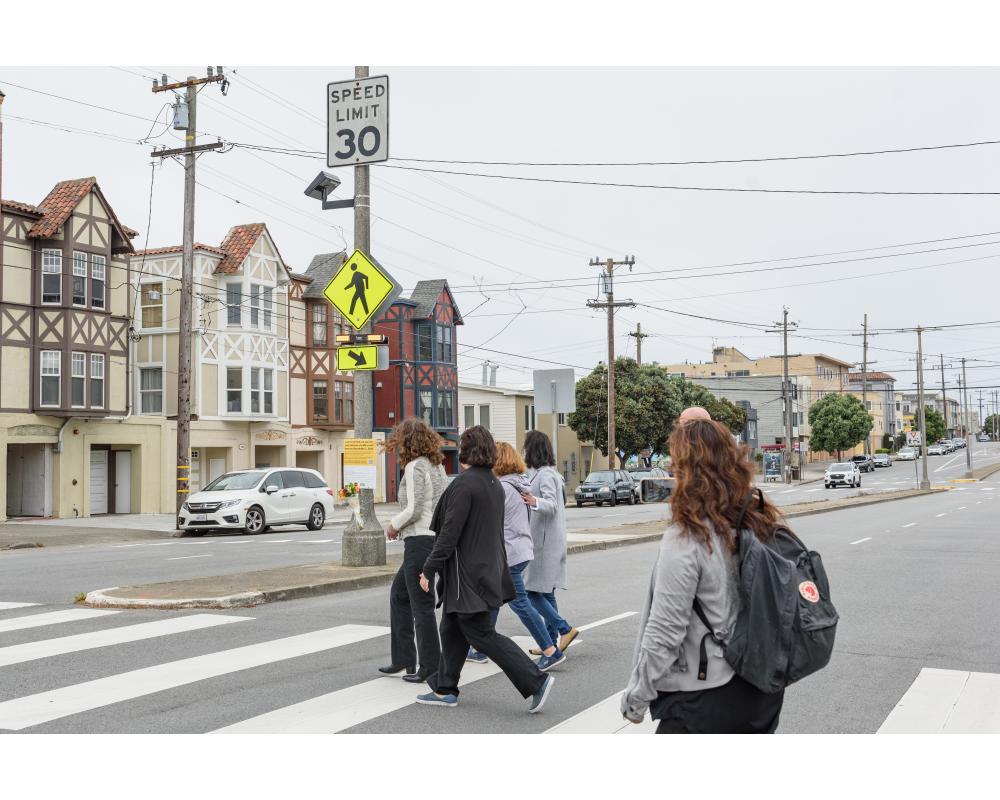 Several people cross a street near a yellow rapid flashing beacon. It includes a yellow sign and one with an arrow.