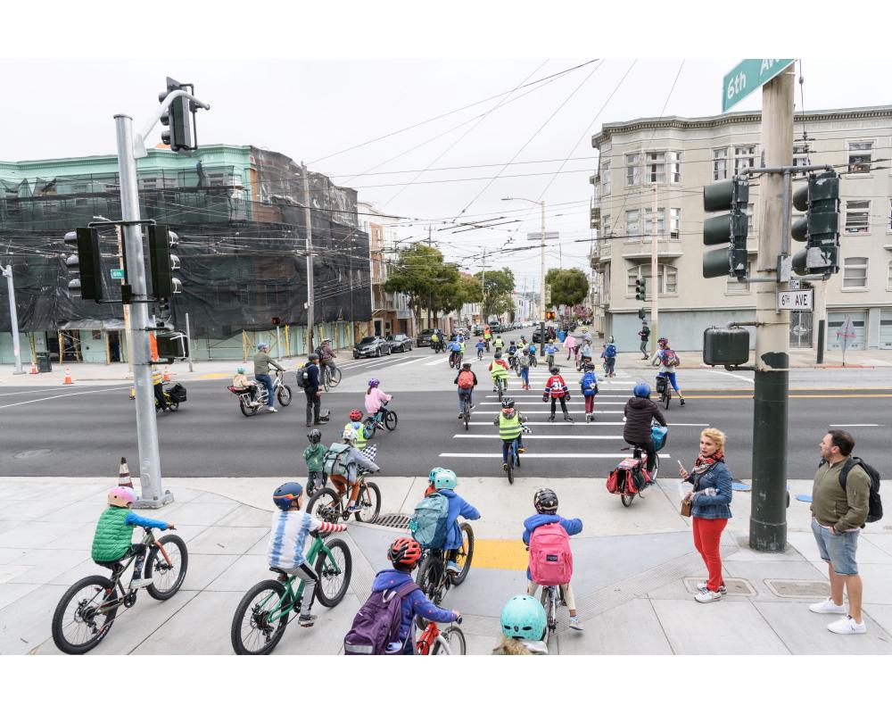 Students wearing helmets and safety vests bike across a street during a school event.