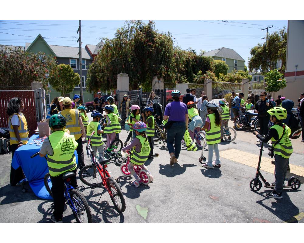 Students wearing yellow vests pause with their bikes by a table with staff during a school event.