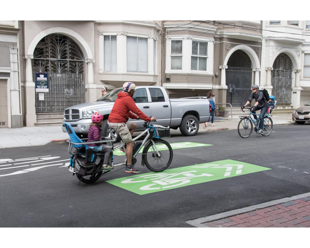 An adult and child ride a two-person bike on a bikeway that has green paint indicating bikes will be on the roadway.