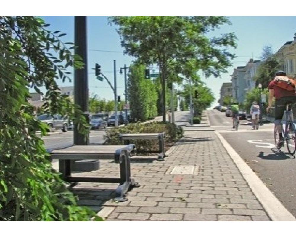 A person bikes near a paved area that has benches and green trees.
